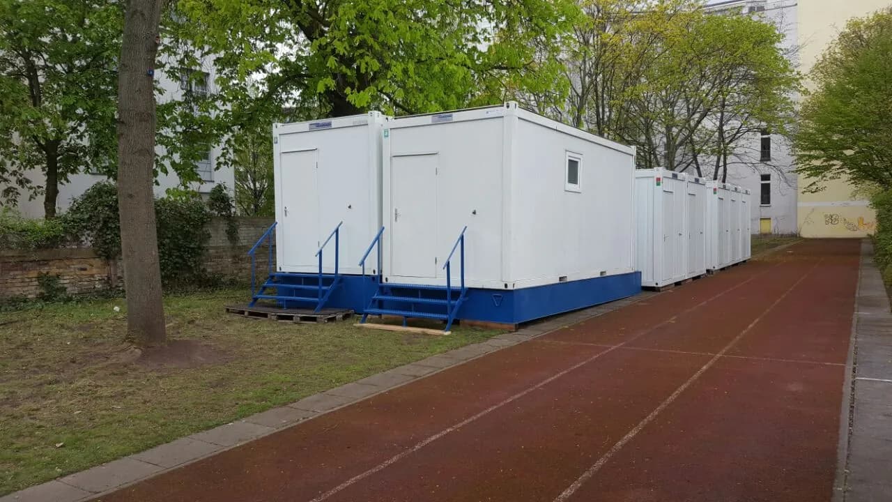 Mehrere Sanitärcontainer mit blauem Sockel und Zugangstreppen stehen auf einem Schulhof – ideal als temporäre WC- oder Duschlösung bei Veranstaltungen oder Baustellen.